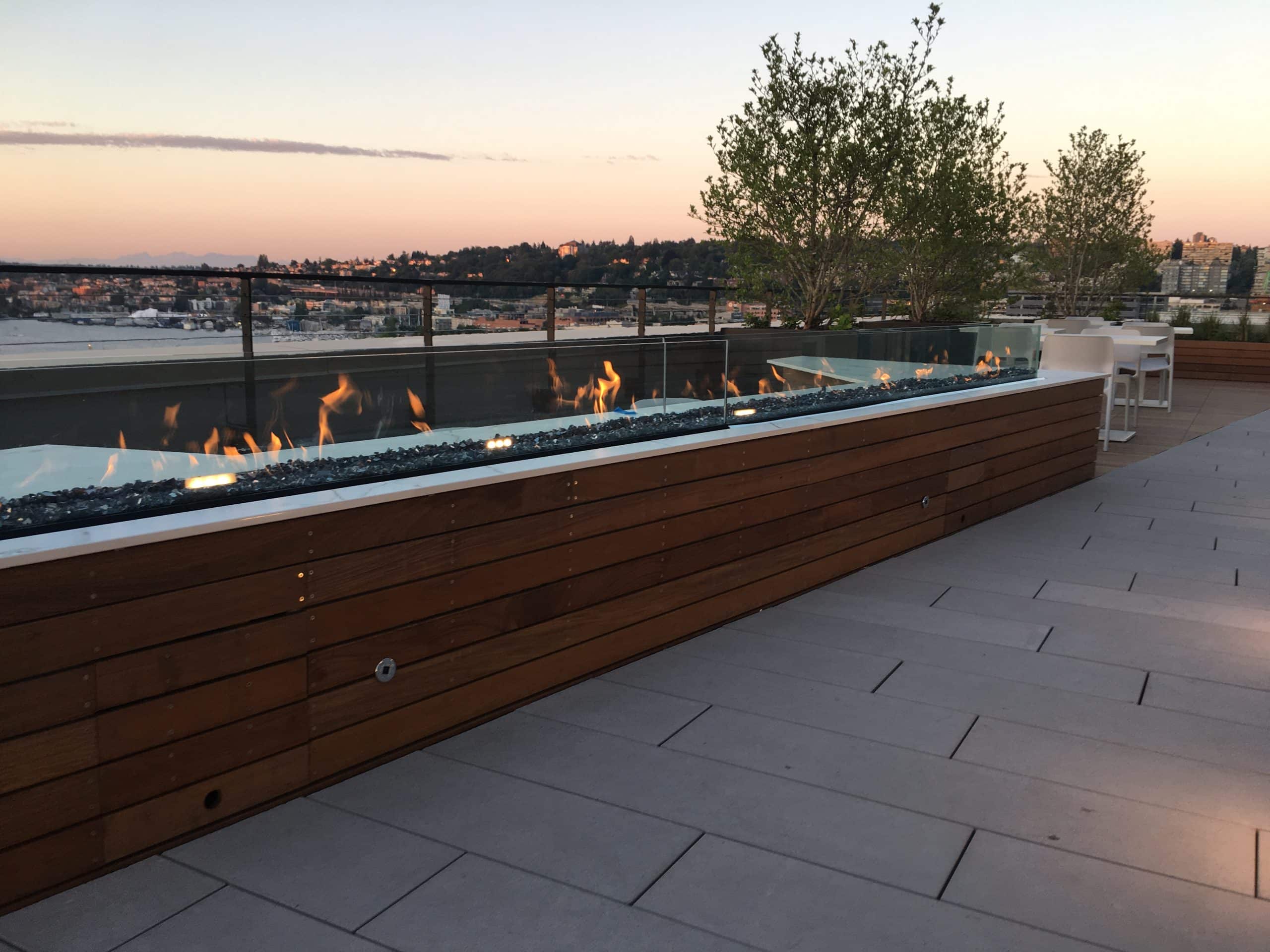 A modern rooftop terrace featuring a sleek linear gas fireplace with wind screens and glass media.